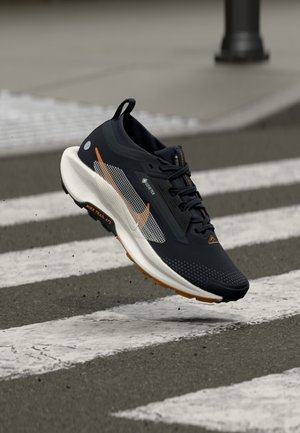 Chaussure de trail Nike noire et blanche avec des détails orange flottant au-dessus d'un passage piéton sur une route asphaltée.