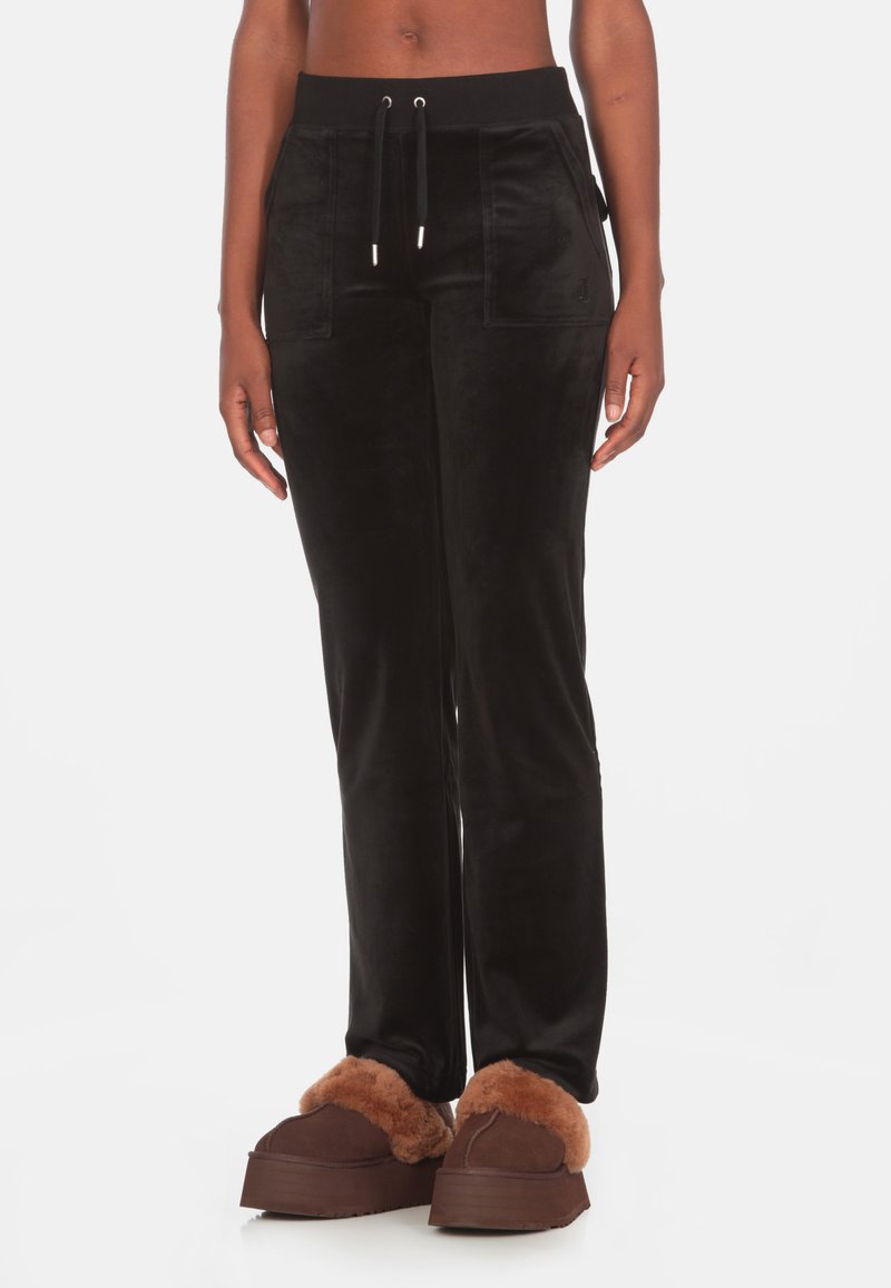 Jogging en velours noir avec une taille élastique à cordon de serrage, des poches latérales et une coupe ajustée, associé à des chaussures d'intérieur en fourrure marron.