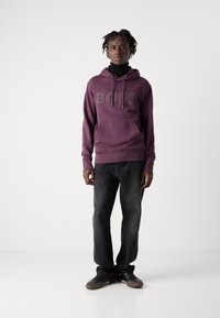 Lila Kapuzenpullover mit "BOSS"-Logo, Fronttasche, Kordelzugkapuze, kombiniert mit dunkelgrauen Jeans und schwarzen Sneakers. Glatter Stoff und lockere Passform.