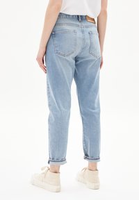 Personne portant un jean taille haute bleu clair avec les revers retroussés et des baskets beige à lacets, debout contre un fond blanc, vue de dos.
