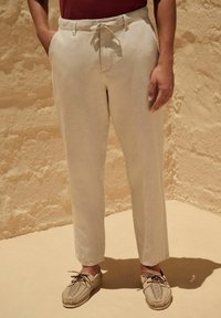 Beige linnen broek met een trekkoord in de taille, voorzien van zijzakken en een relaxte pasvorm, gecombineerd met lichtbeige gevlochten schoenen.