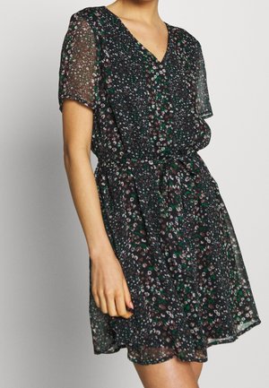 Kobieta ubrana w czarną sukienkę w kwiaty z krótkim rękawem, dekoltem w serek i paskiem zawiązanym w talii, stojąca na jednolitym tle.