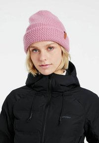 Rosa Strickmütze mit einem strukturierten Muster und einem Leder-Logo-Patch, getragen zu einer schwarzen Kapuzenjacke mit Reißverschluss.
