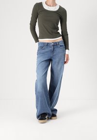 Jeans blu larghi e lunghi con un lavaggio sbiadito, abbinati a una maglietta aderente verde oliva con bordi bianchi e un design corto.