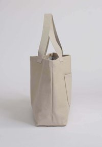 Bolsa tote de lona beige con dos asas de tela, bolsillo lateral y forro interior a rayas. Forma rectangular y resistente con una textura suave.