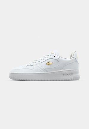 Sneaker in pelle bianca con superficie liscia, punta tondeggiante e chiusura con lacci. Presenta il logo dorato di Lacoste sul lato e sul tallone. Suola in gomma.