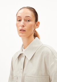 Hellgraue Jacke mit spitzem Kragen, zwei vorderen Taschen und Knopfverschluss. Der Stoff hat eine subtile Textur.