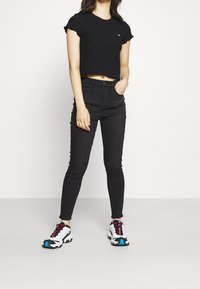 Woman wearing a black cropped top, high-waisted black skinny jeans, and white sneakers with blue and red accents, standing with one hand in pocket.