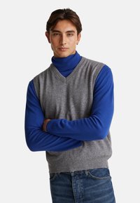Męski sweter bez rękawów w szarym wełnianym kolorze na niebieskiej bluzce z golfem. Posiada dekolt w serek i żebrowane mankiety, noszony z dżinsami.