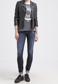 Giubbotto da biker in pelle nera, t-shirt grafica grigia, jeans skinny in denim scuro e sneakers nere con lacci. Outfit urbano casual.