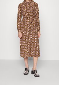 Långärmad klänning i brunt med ett geometriskt mönster i lila, kräm och grönt. Inkluderar en bälte i midjan och en krage. Matchas med blanka svarta brogues.