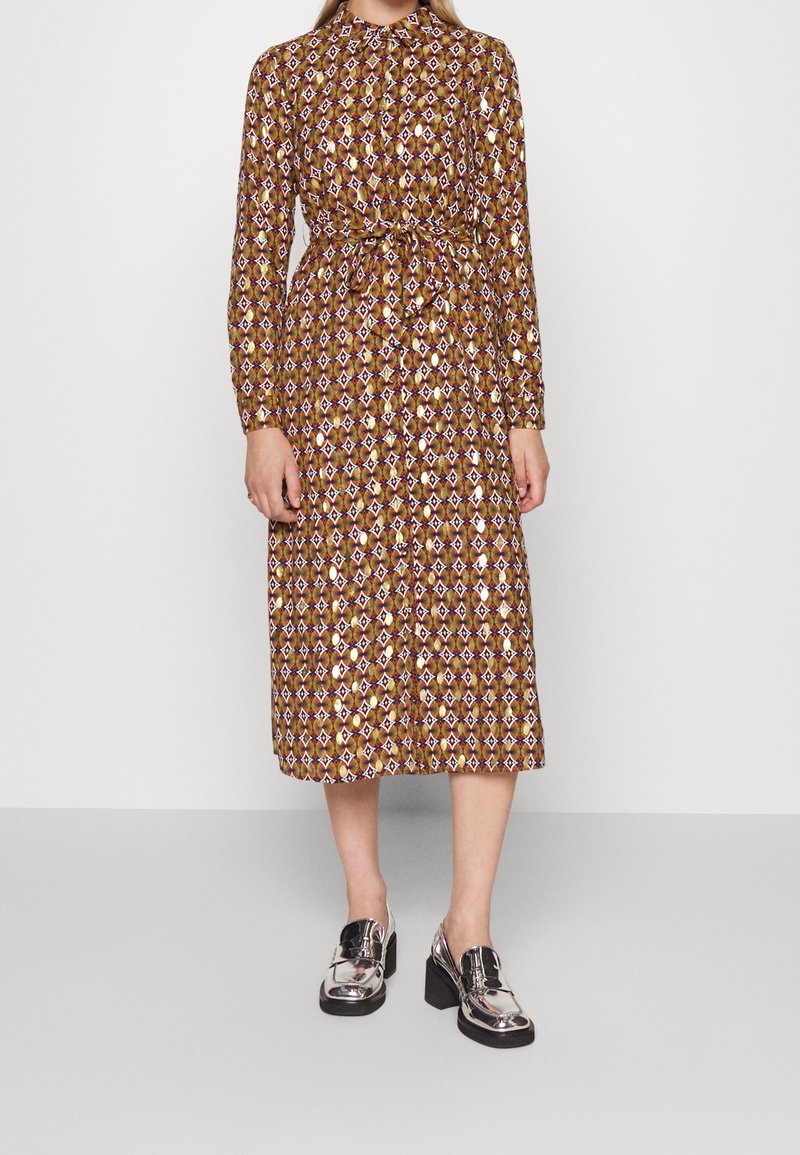 Långärmad klänning i brunt med ett geometriskt mönster i lila, kräm och grönt. Inkluderar en bälte i midjan och en krage. Matchas med blanka svarta brogues.