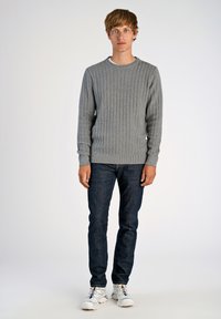 Szary sweter w warkoczowy splot z okrągłym dekoltem, w połączeniu z ciemnoniebieskimi dżinsami i białymi trampkami. Model stoi na jednolitym tle.