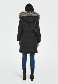 Sort isoleret jakke med en hætte foret med pels, der har chevron-syninger og ribbet tekstur på siderne, parret med blå jeans og sorte støvler.