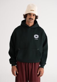 Felpa oversize verde scuro con tasca frontale e logo bianco, abbinata a pantaloni bordeaux. Materiale in cotone, texture morbida, design minimale.