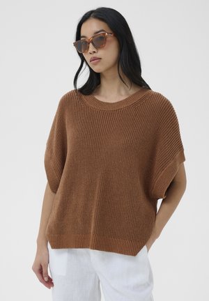 Femme aux longs cheveux noirs portant des lunettes de soleil écaille de tortue, un pull en maille marron ample à manches courtes et un pantalon blanc sur un fond uni.