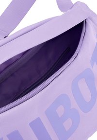 Fioletowa torba wykonana z tkaniny, z kieszenią wewnętrzną na zamek błyskawiczny. Gładka tekstura z dużym nadrukiem logo na zewnątrz.