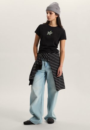 Black t-shirt with green star graphic, light blue wide-leg jeans, black and white striped sweater tied around waist, and gray beanie.