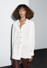 Bílá košile na knoflíky s velkým límcem, ozdobená zlatými knoflíky a volnějším střihem. Nošena s hnědými koženými kalhotami, na šedém pozadí.