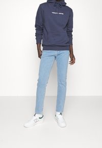 Marinblå hoodie med "Tommy Jeans" logotyp, ljusblå denimjeans och vita sneakers med gröna detaljer. Slät tyg och figurnära design.