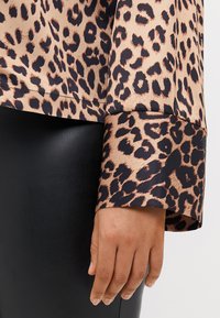 Blusa com estampa de leopardo e acabamento acetinado. Apresenta punhos largas em preto, textura suave e bainha arredondada. Combinada com leggings pretas.
