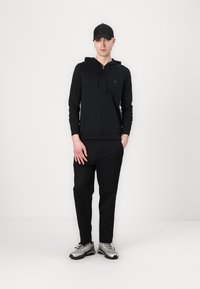 Felpa nera con zip e cappuccio regolabile, abbinata a pantaloni neri. Presenta un logo minimalista ed è completata da sneaker argento.