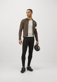 Brown zip-up cycling jacket over a white base layer, paired with black leggings and cycling shoes; holding a black helmet.