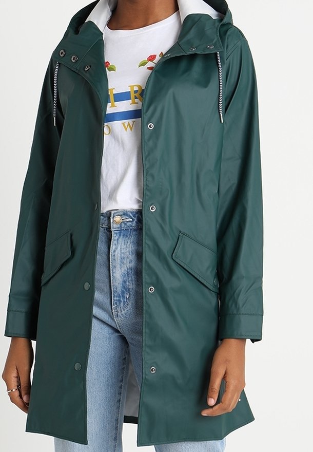 Imperméable à capuche vert foncé avec des boutons-pression à l'avant porté sur un t-shirt blanc à motifs et un jean bleu clair sur une personne debout.