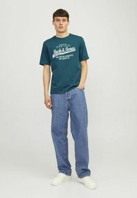 Teal bomulds t-shirt med hvid grafisk tekst, parret med løstsiddende blå denimjeans og hvide sneakers. Simpelt design og afslappet pasform.