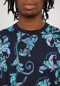 Temno modra majica s čudovitimi svetlo modrimi cvetnimi vzorci po celotni površini, z teksturiranimi detajli in integriranim besedilom "Versace Jeans Couture".