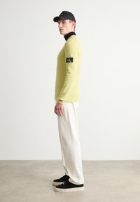 Lys grønn genser med svart lapp, parret med hvite vide bukser og svarte sneakers med tykk såle, stylet med en svart caps.