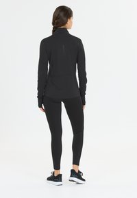 Zwart sporttop met een hoge kraag en lange mouwen, gecombineerd met zwarte leggings en zwarte sneakers, die een slank ontwerp benadrukken.
