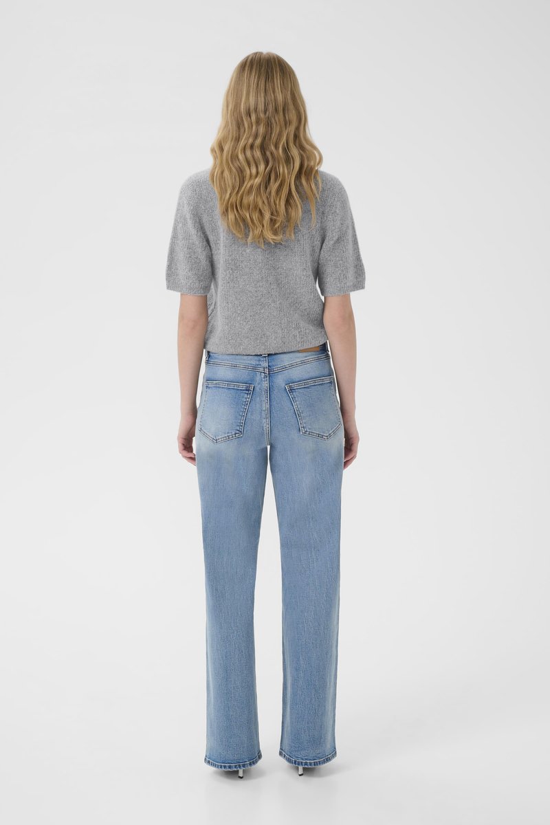 Woman with wavy blonde hair wearing a gray short-sleeve sweater and light blue straight-leg jeans, standing facing away on a white background.