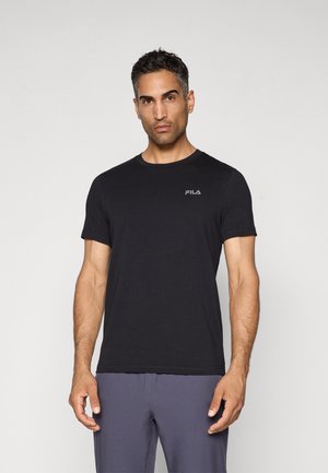 Mand med kort mørkt hår iført en sort FILA t-shirt og grå bukser, stående foran en ensfarvet hvid baggrund.