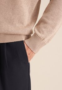 Beige gestrickter Pullover mit gerippten Bündchen, getragen über dunklen Hosen, der eine weiche Textur und eine taillierte Form zeigt. Schlicht ohne sichtbare Muster.