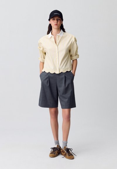 Camicia a righe gialle e bianche con bottoni, pantaloni corti grigi plissettati, calze grigie, scarpe sportive marroni, cappellino nero con logo. Orlo ondulato sulla camicia.