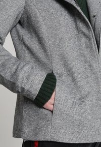 Grijze jas met textuurstof, een kraag en groene geribbelde accenten op de manchetten, met een zichtbare zijzak.