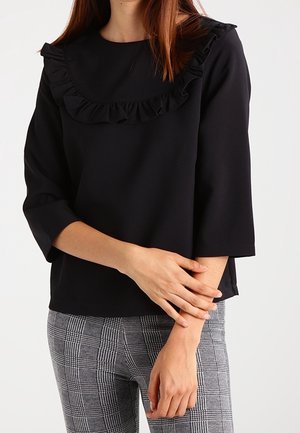 Kobieta ubrana w czarną bluzkę z długim rękawem i marszczonym dekoltem oraz szare spodnie w kratę, pozująca z rękami skrzyżowanymi na wysokości talii.