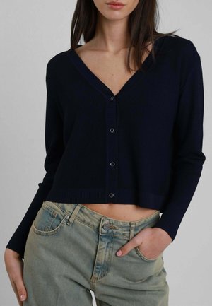 Giovane donna che indossa un cardigan corto blu navy con bottoni e jeans a vita alta chiari, con una mano in tasca.