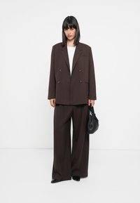 Sacou maro cu două rânduri de nasturi și rever larg, asortat cu pantaloni largi. Accesorii includ o geantă mică neagră și pantofi negri cu vârful ascuțit.