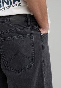 Mustat farkut, joissa on takatasku, jossa on erottuva tikkauskuvio ja suorakaiteen muotoinen nahkalappu vyötärövyössä.