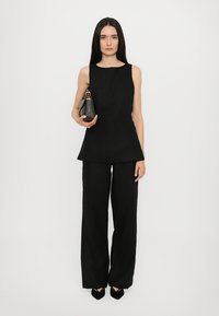 Svart ärmlös topp med peplumsläpp, kombinerad med vida svarta byxor. Modellen håller en liten svart väska med guldaccenter. Slät textur.