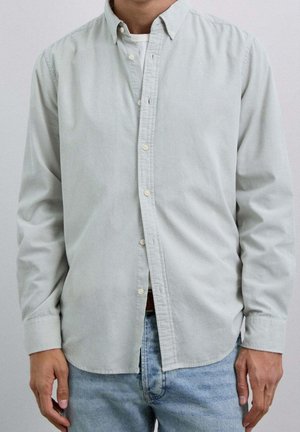 Mand iført en lysegrå skjorte med knapper over en hvid t-shirt og lyseblå jeans, stående med armene afslappet langs siden.