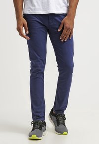 Navy blue slim-fit pants made of cotton blend, featuring standard pockets and a smooth texture, paired with black and gray sneakers with yellow accents.