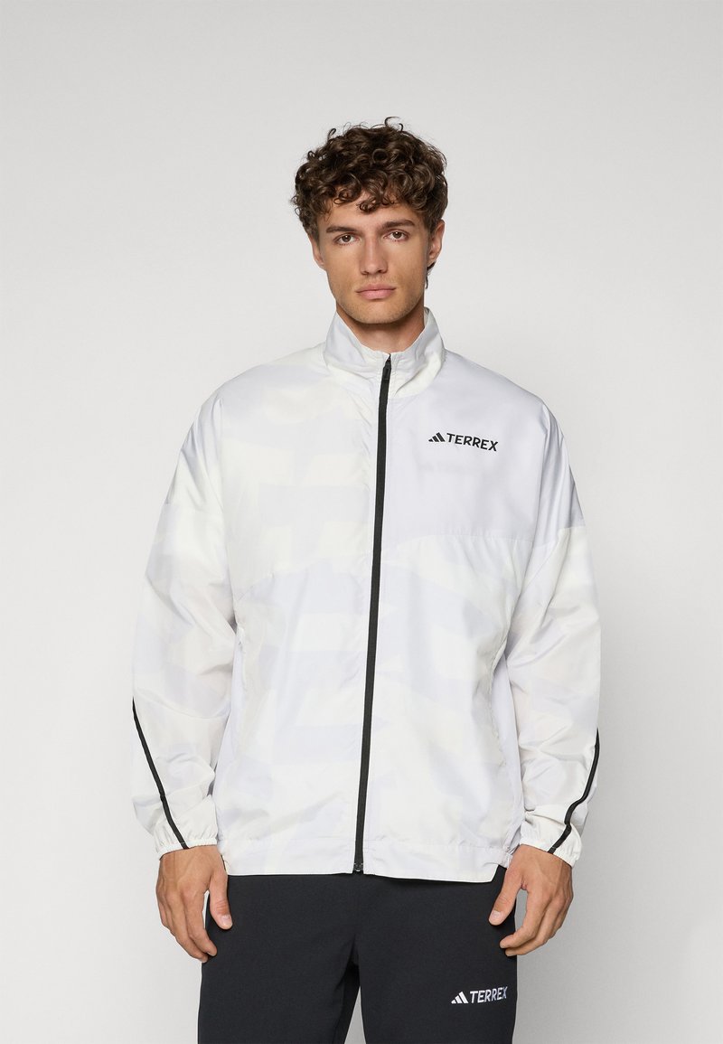 Veste Terrex blanche avec col montant, fermeture éclair à l'avant et poignets élastiques. Présente des accents noirs et un motif géométrique subtil.