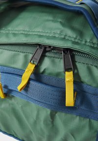 Backpack with a green textured fabric, featuring yellow zip pulls, blue accents, and YKK zipper hardware. Zip compartment visible at top.