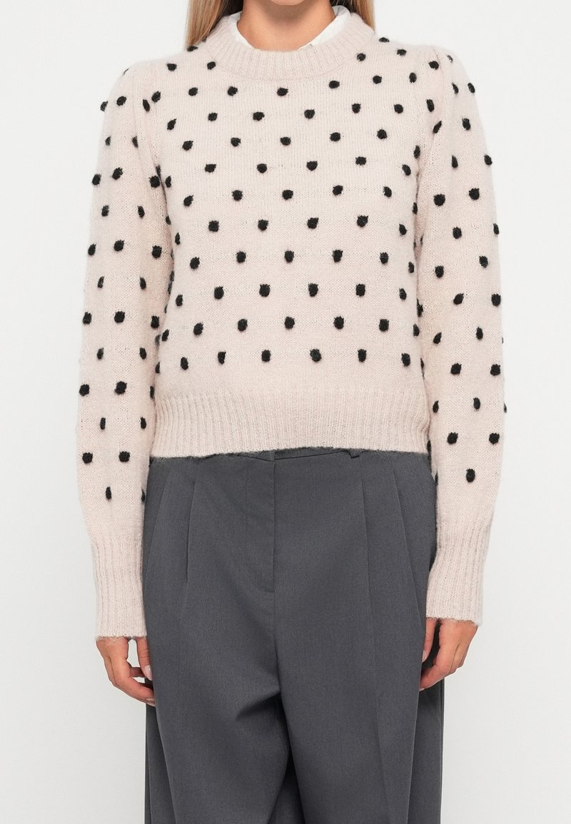 Maglione rosa chiaro con pois a pallini neri, collo e orlo a coste. Indossato con pantaloni grigi a vita alta, con dettagli a pieghe.