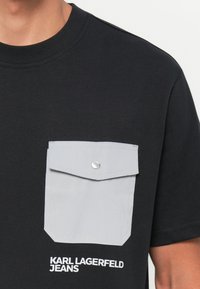 Juodas marškinėliai su pilku kišenės pleistru, sidabriniu spaudimo uždarymu ir balta "KARL LAGERFELD JEANS" užrašu priekyje. Lygios medžiagos tekstūra.