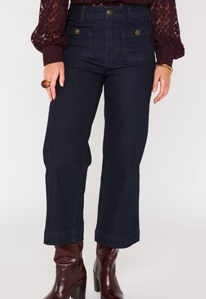 Pantaloni a gamba larga in denim scuro con tasche frontali, dettagli con bottoni dorati e orlo risvoltato. Abbinati a stivali con tacco lucidi bordeaux.