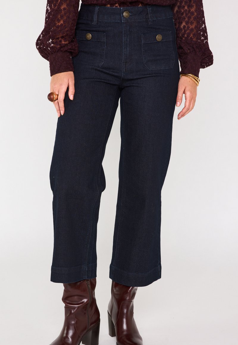 Pantaloni a gamba larga in denim scuro con tasche frontali, dettagli con bottoni dorati e orlo risvoltato. Abbinati a stivali con tacco lucidi bordeaux.